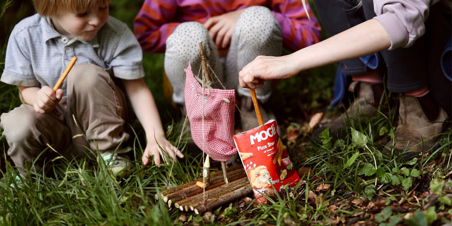 Kinder spielen draußen mit einem kleinen Floß aus Stöcken, Stoffsegel und Snacks im Gras