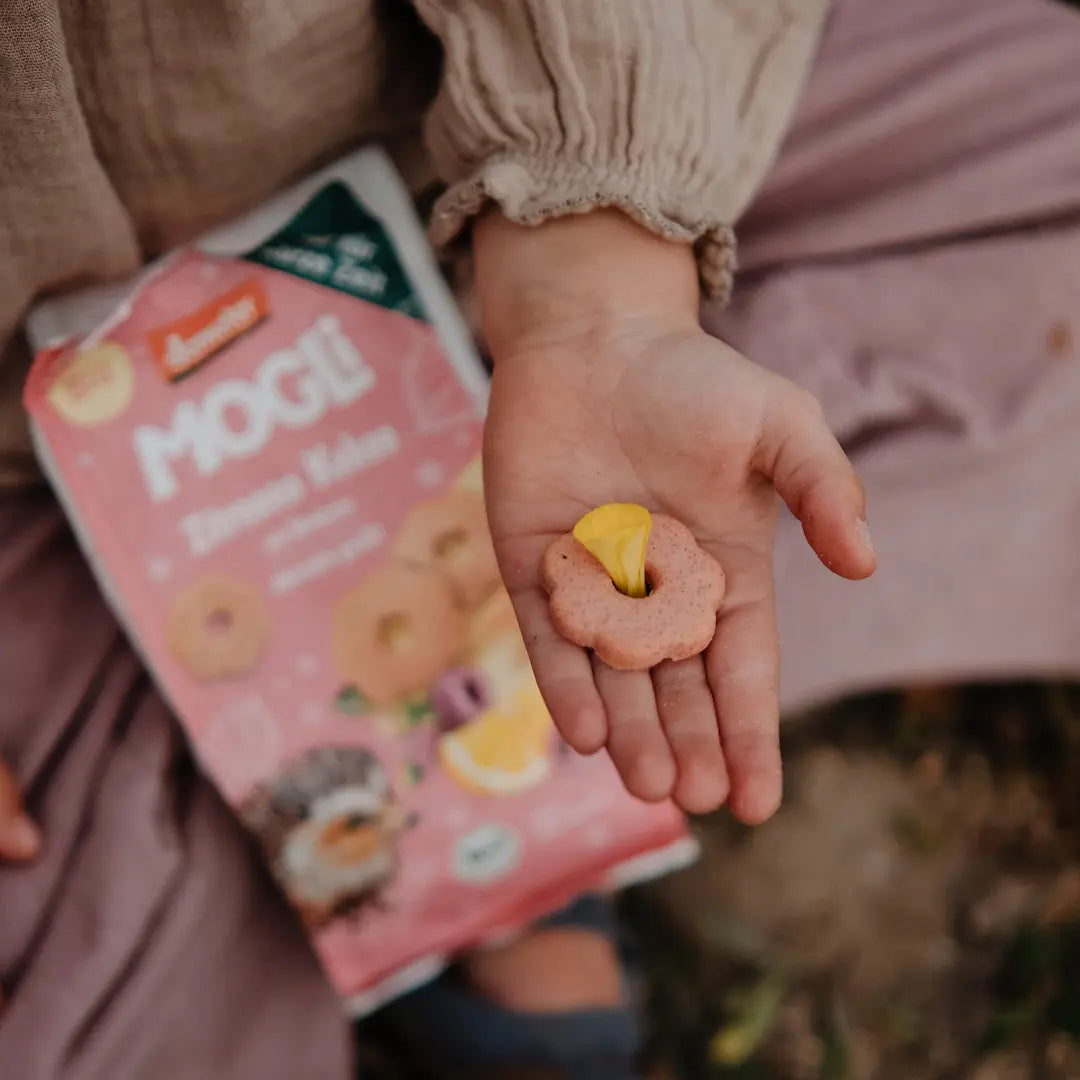 Eine Kinderhand hält einen MOGLi Zitronen Himbeer Keks in der Hand, in der Mitte vom Keks ist eine gelbe Blüte und unter der Hand liegt die ausgepackte Verpackung von den Keksen.