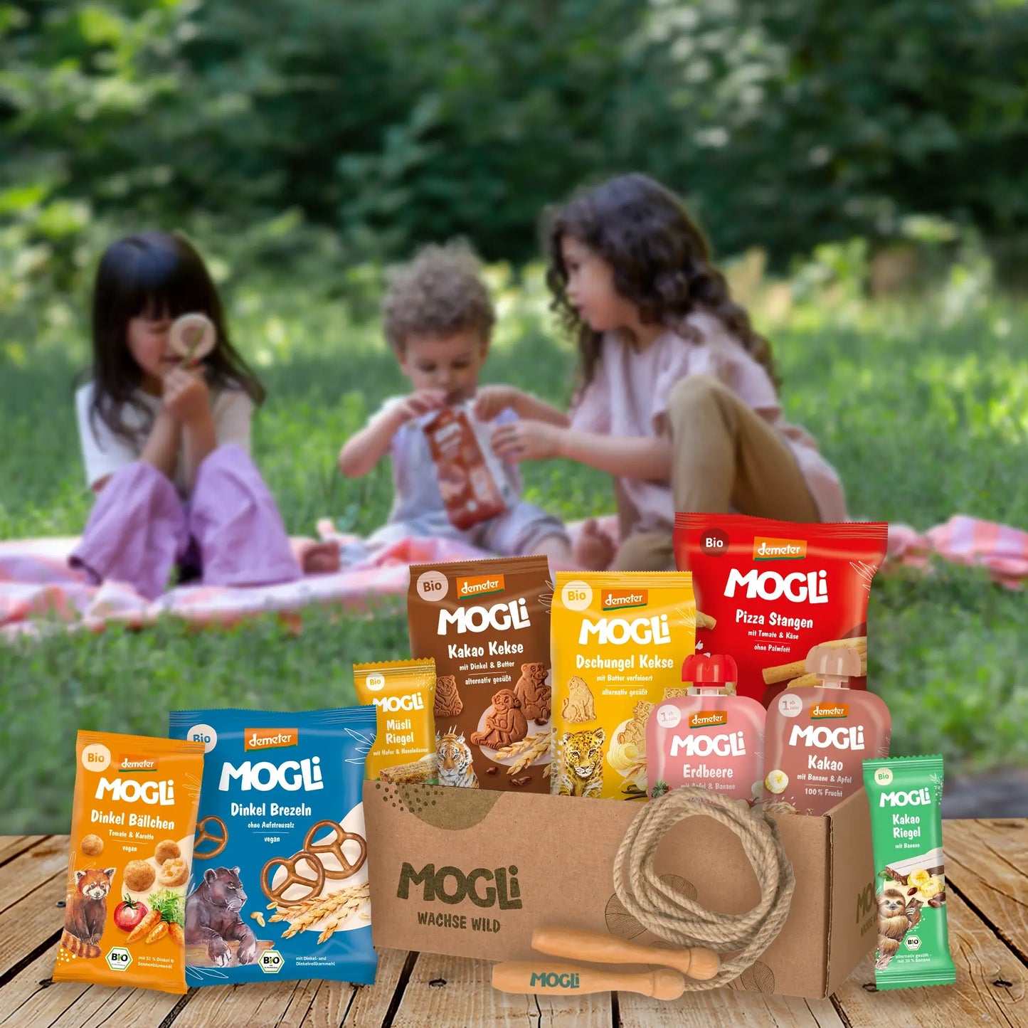 MOGLi Ausflugs-Bundle mit Bio- und Demeter-Snacks und Springseil auf Holzboden; im Hintergrund picknicken drei Kinder im Grünen.