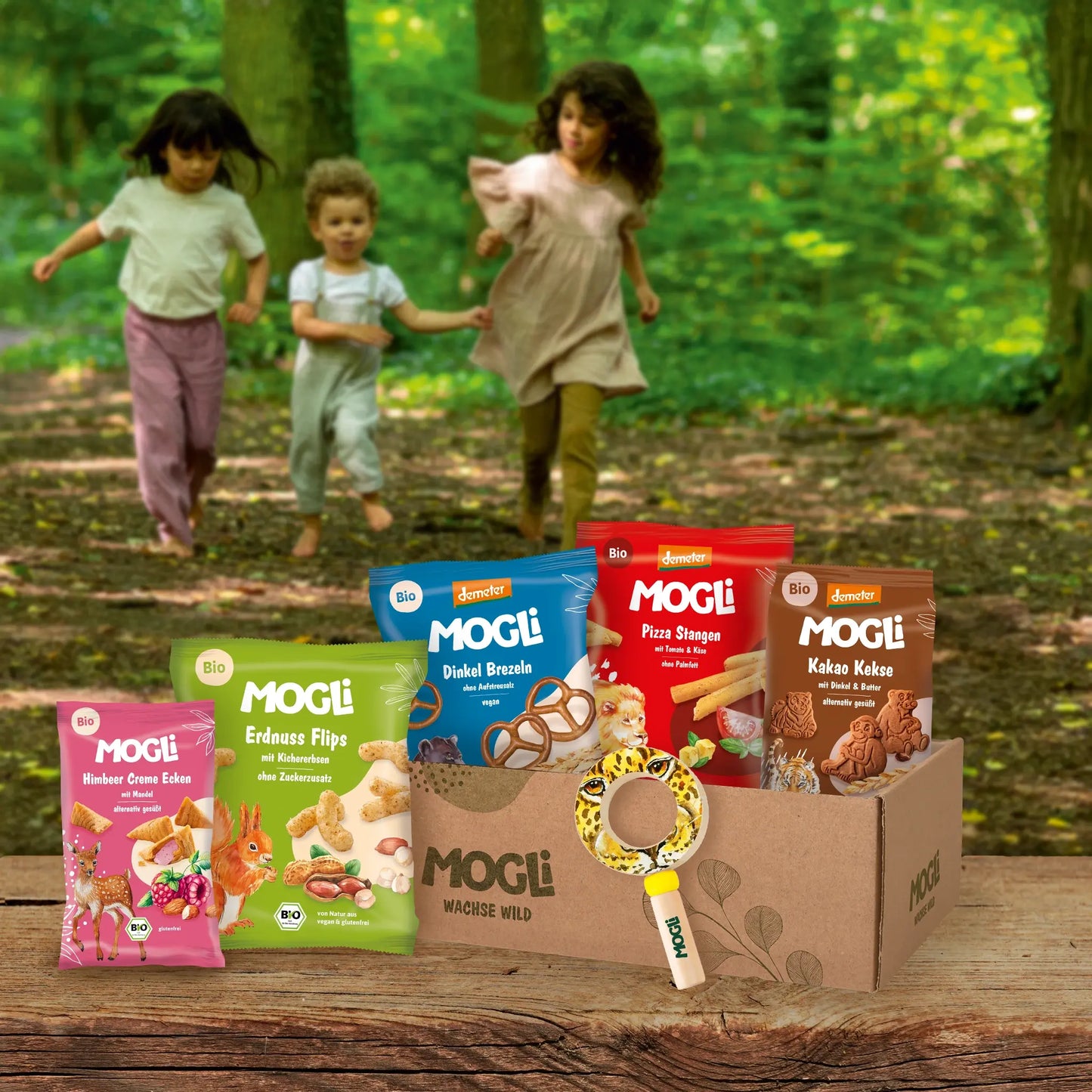 MOGLi Erlebnis-Bundle mit Bio- und Demeter-Snacks und Kinderlupe auf Holzboden; im Hintergrund rennen drei Kinder durch den Wald.