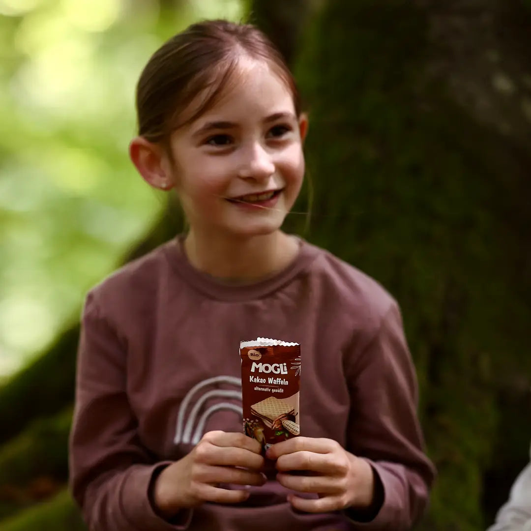 Kind hält verpackte MOGLi Kakao Waffel in der Hand, sitzt im Wald und lächelt.