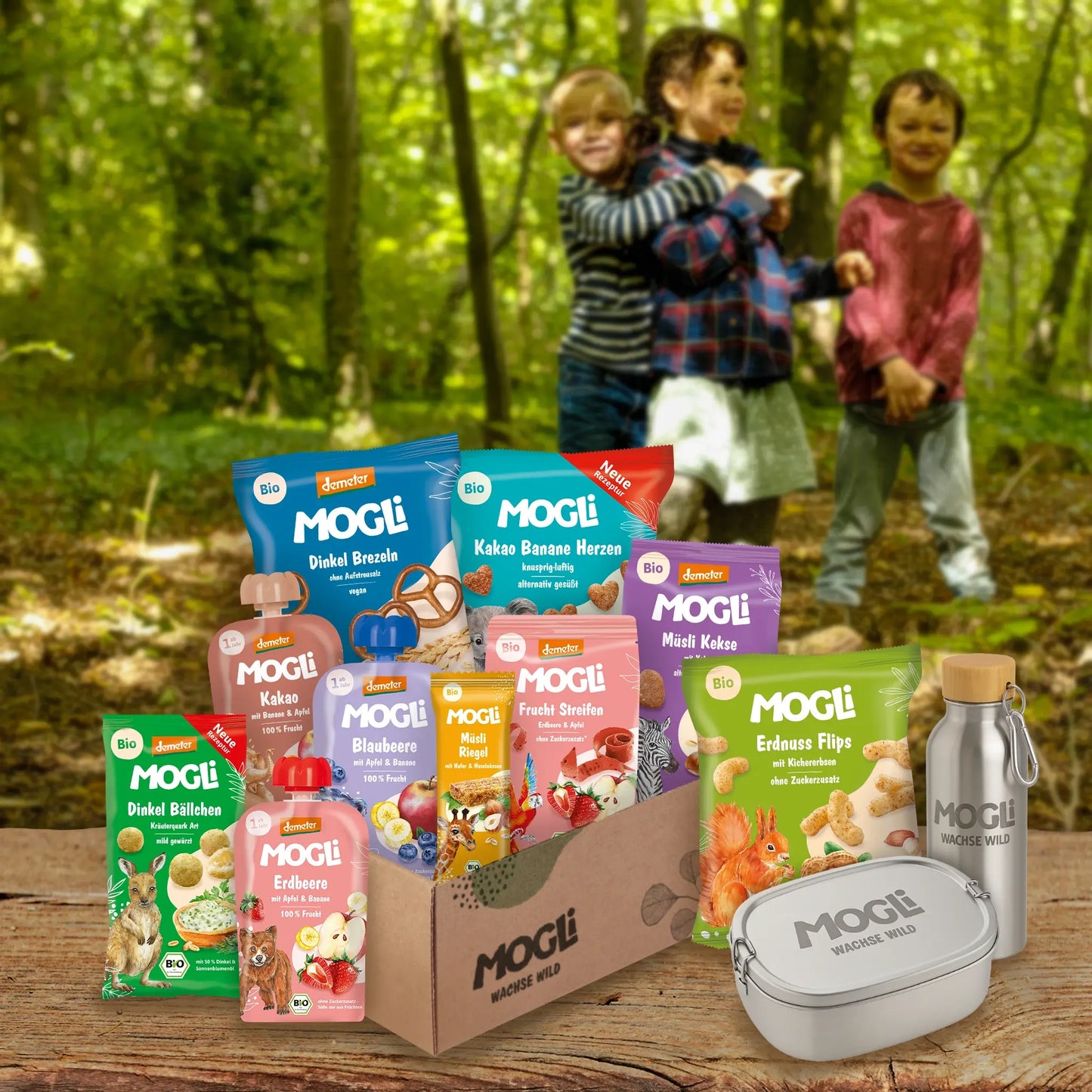 MOGLi Kita Knabber Bundle mit Bio- und Demeter-Snacks, Brotdose und Trinkflasche vor Waldkulisse; im Hintergrund spielen drei Kinder zwischen den Bäumen im Wald.