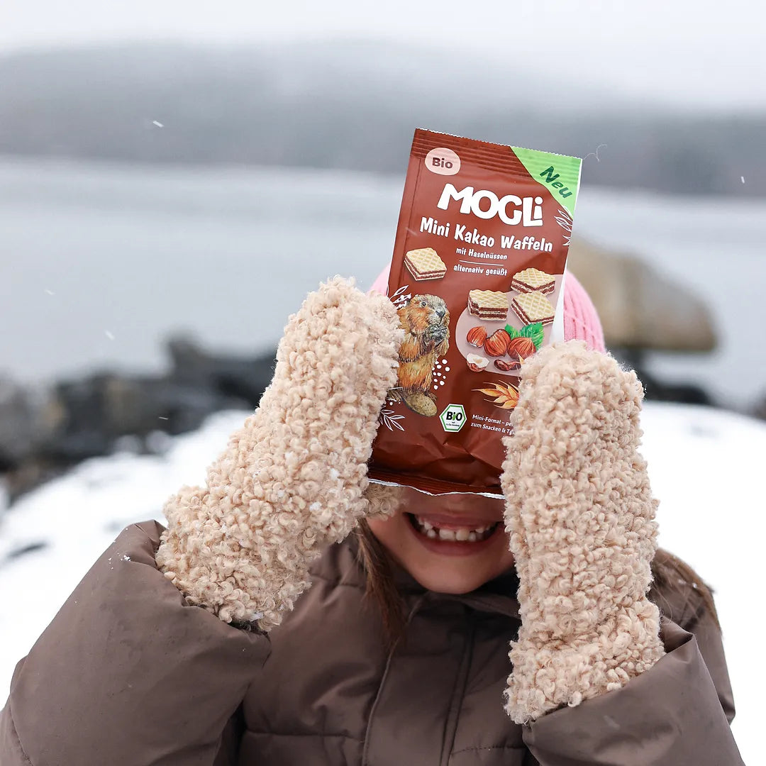 Ein Mädchen steht draußen im Schnee und hält eine Packung MOGLi Mini Kakao Waffeln vor ihr Gesicht.