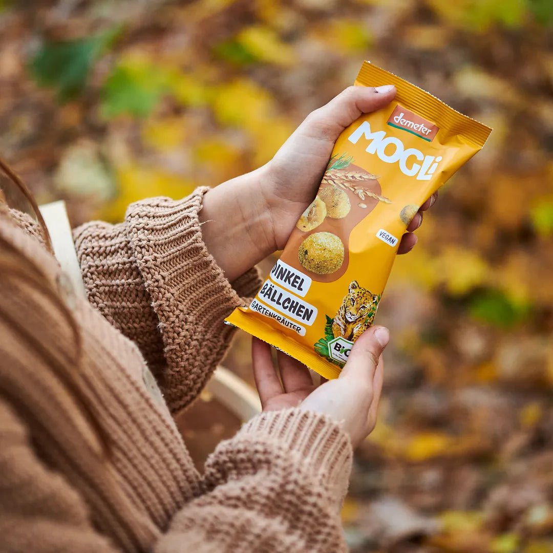 Kind hält eine Packung der MOGLi Dinkel Bällchen in der Hand, im Hintergrund sind verschwommen Herbstblätter