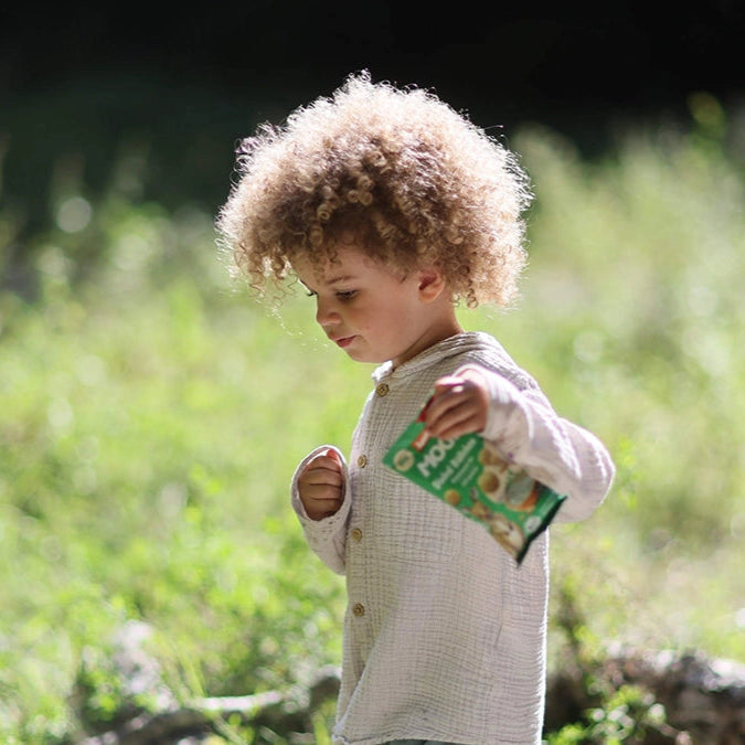 Kleiner Junge läuft über eine Wiese im Wald und hält eine Packung MOGLi Dinkel Bällchen Kräuterquark in der Hand