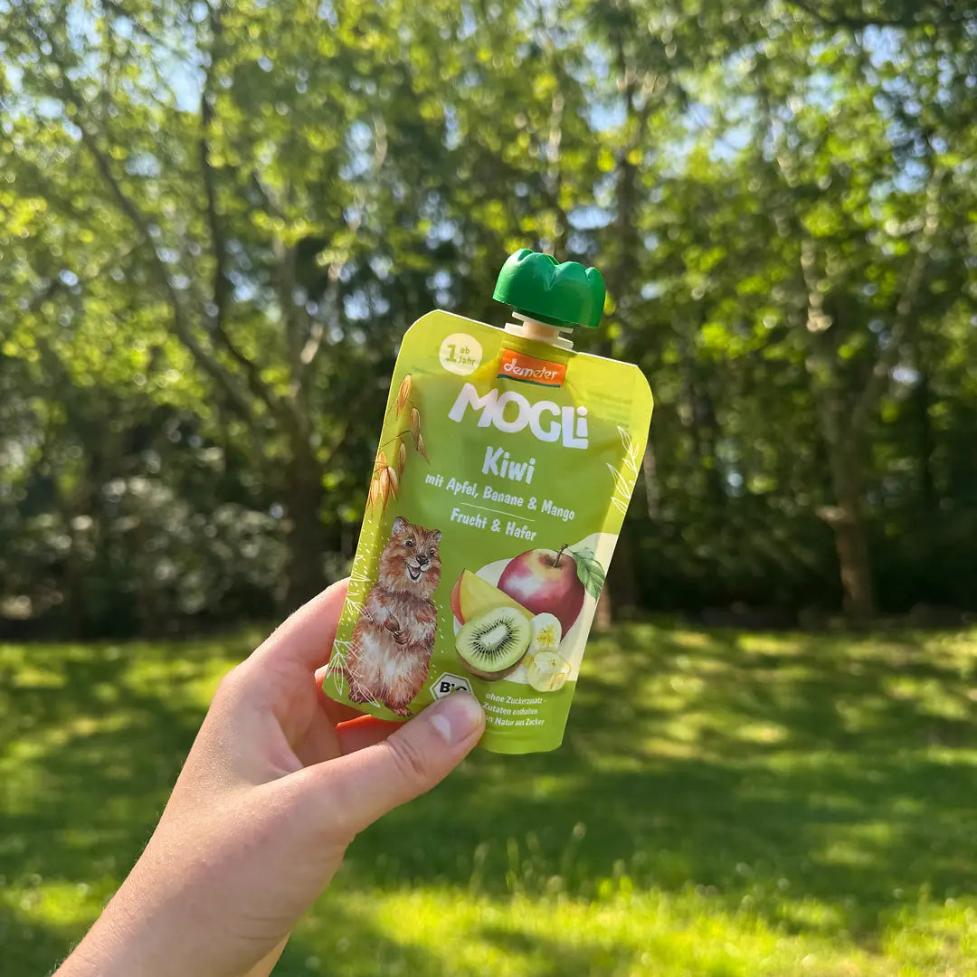 Eine Hand hält in einem Park einen MOGLi Kiwi Quetschie in der Hand. 