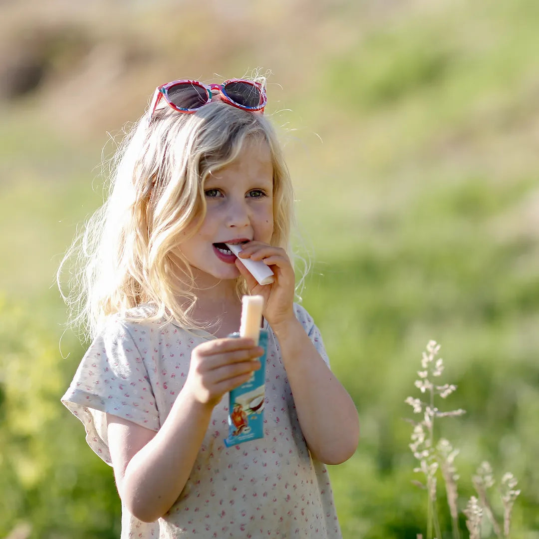 Blondes Mädchen steht auf einem grünen Feld und isst den MOGLi Kokos Riegel