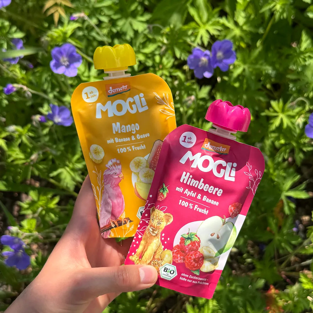 Eine Hand hält einen MOGLi Mango und einen Himbeer Quetschie vor grünen Pflanzen.