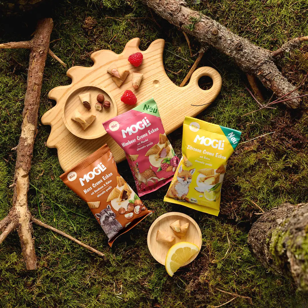 Die MOGLi Creme Ecken Himbeere, Zitrone und Haselnuss liegen auf einem Moosboden und auf einem Holzbrett liegen die ausgepackten Ecken.