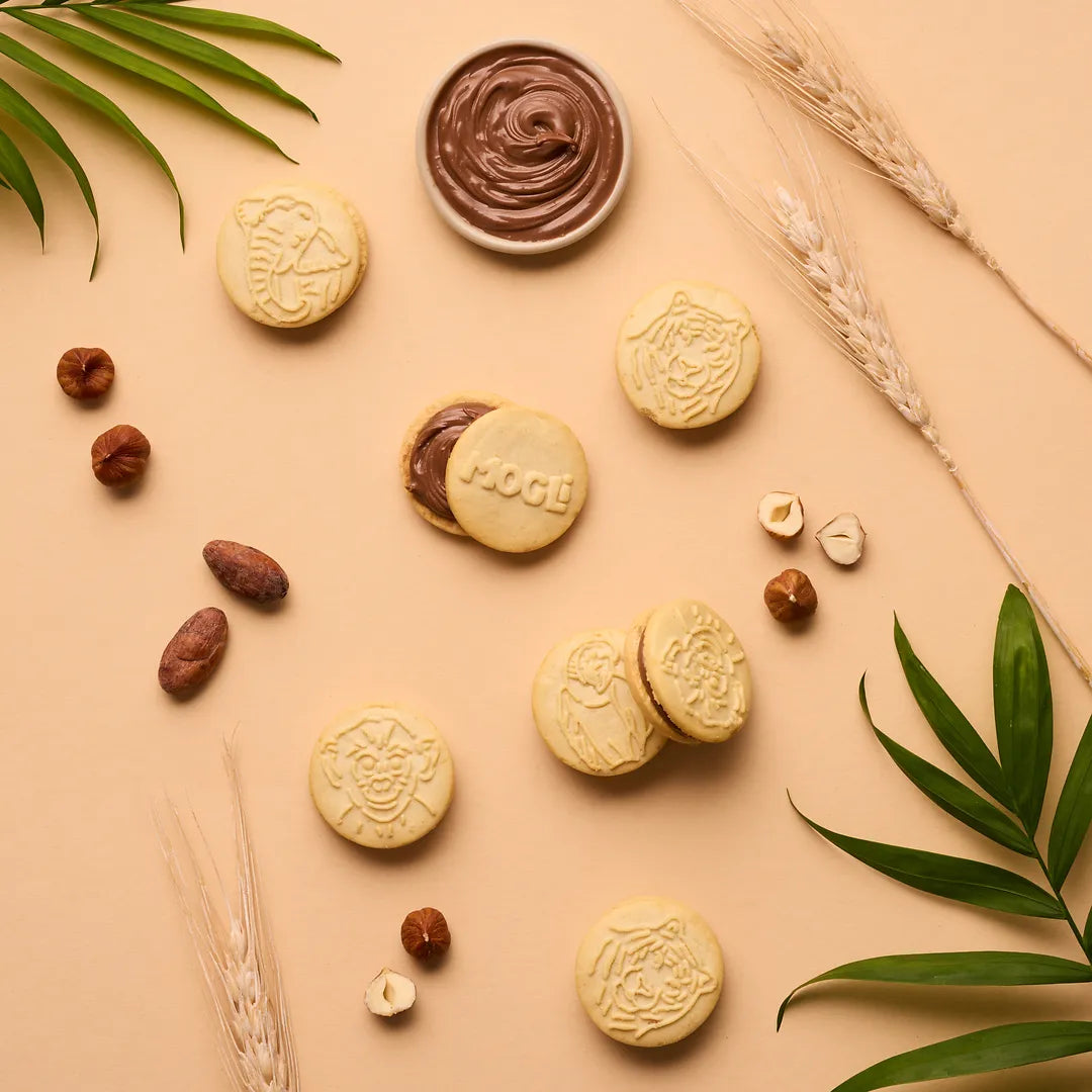 MOGLi Doppel Kekse Kakao mit Haselnuss-Kakao-Creme, umgeben von Haselnüssen, Kakao und Getreide.