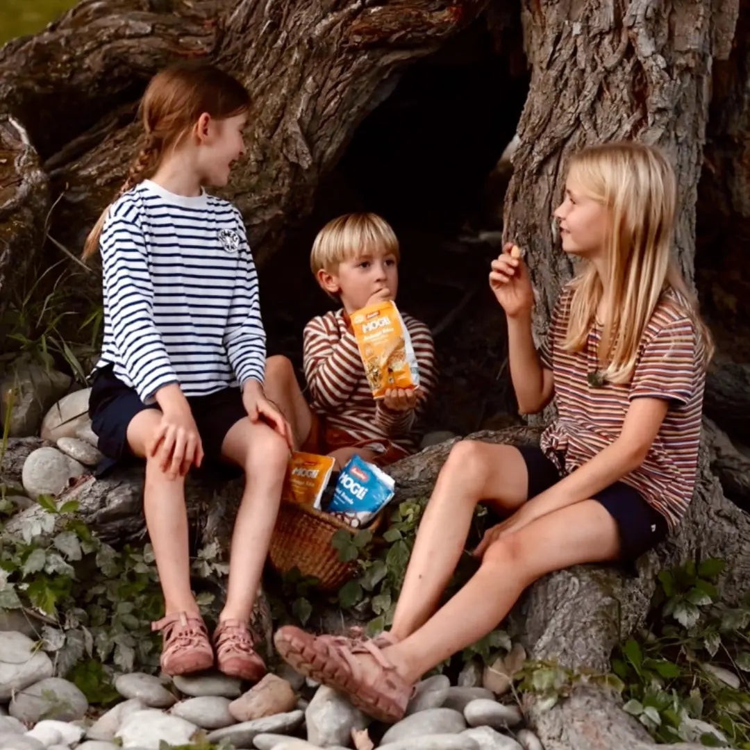 Drei Kinder sitzen im Wald unter einem Baum und teilen sich eine Packung MOGLi Dschungel Kekse.