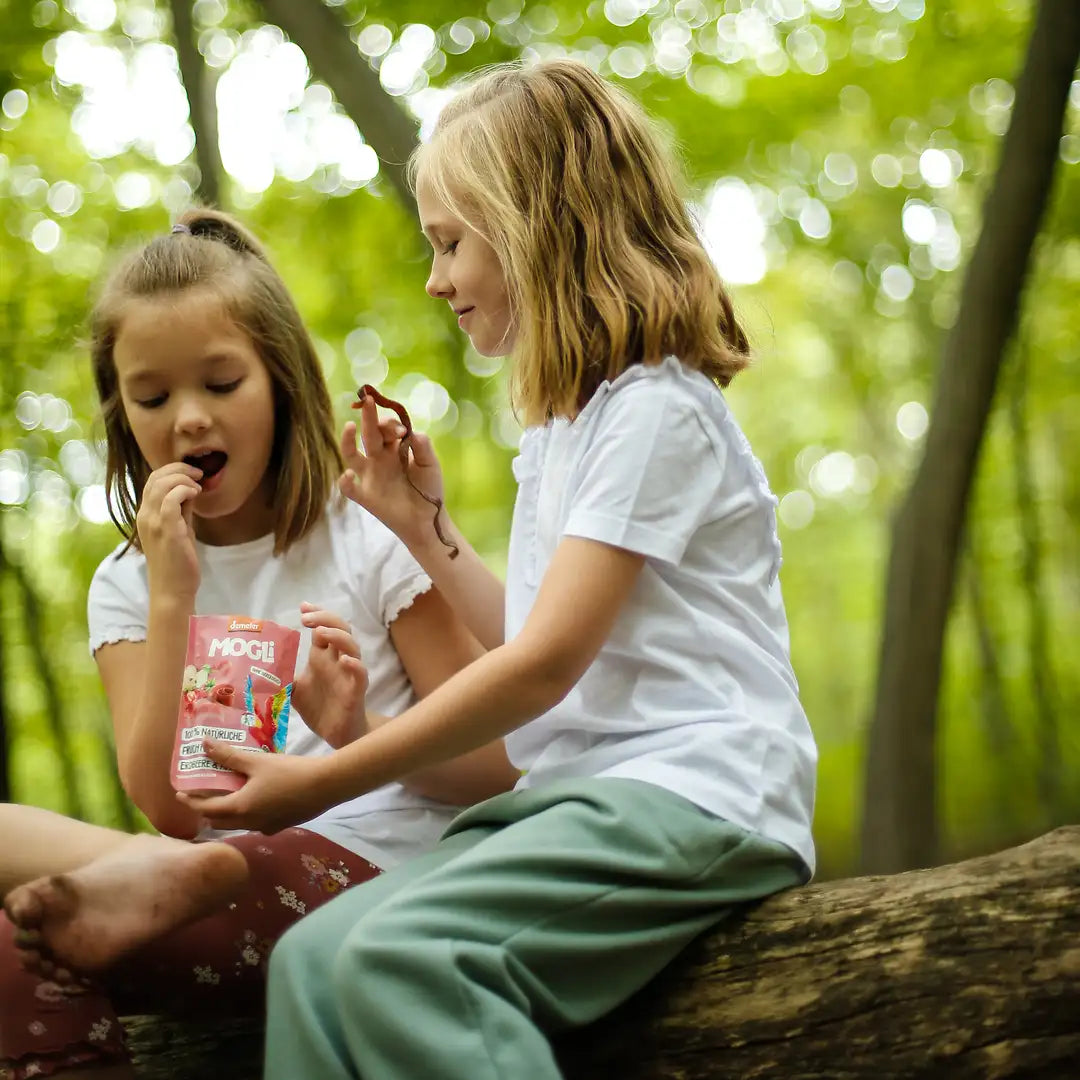 Zwei Mädchen sitzen auf einem Baumstamm im Wald und teilen sich eine Packung MOGLi Erdbeer Frucht Streifen.