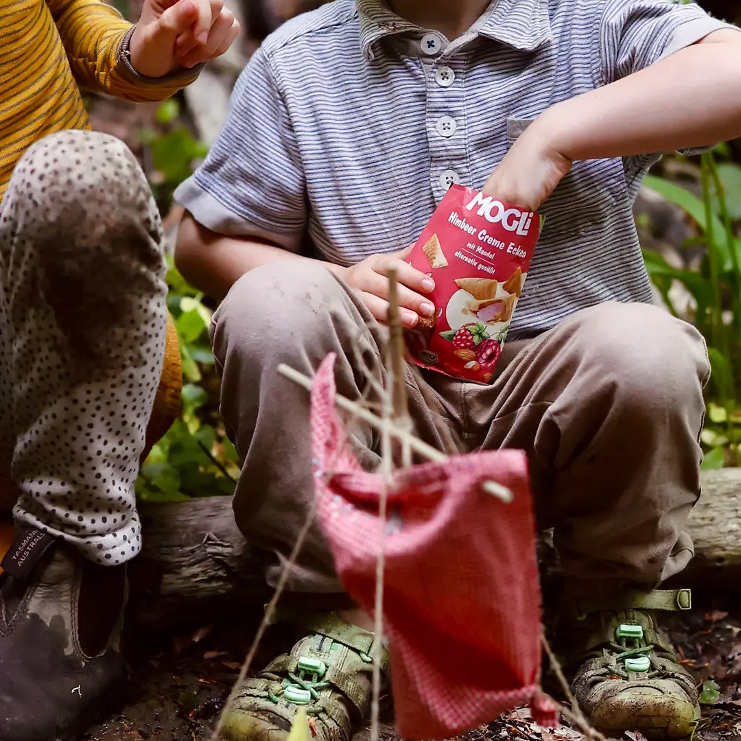 Kind greift in geöffnete Verpackung MOGLi Himbeer Creme Ecken beim Snacken im Wald.