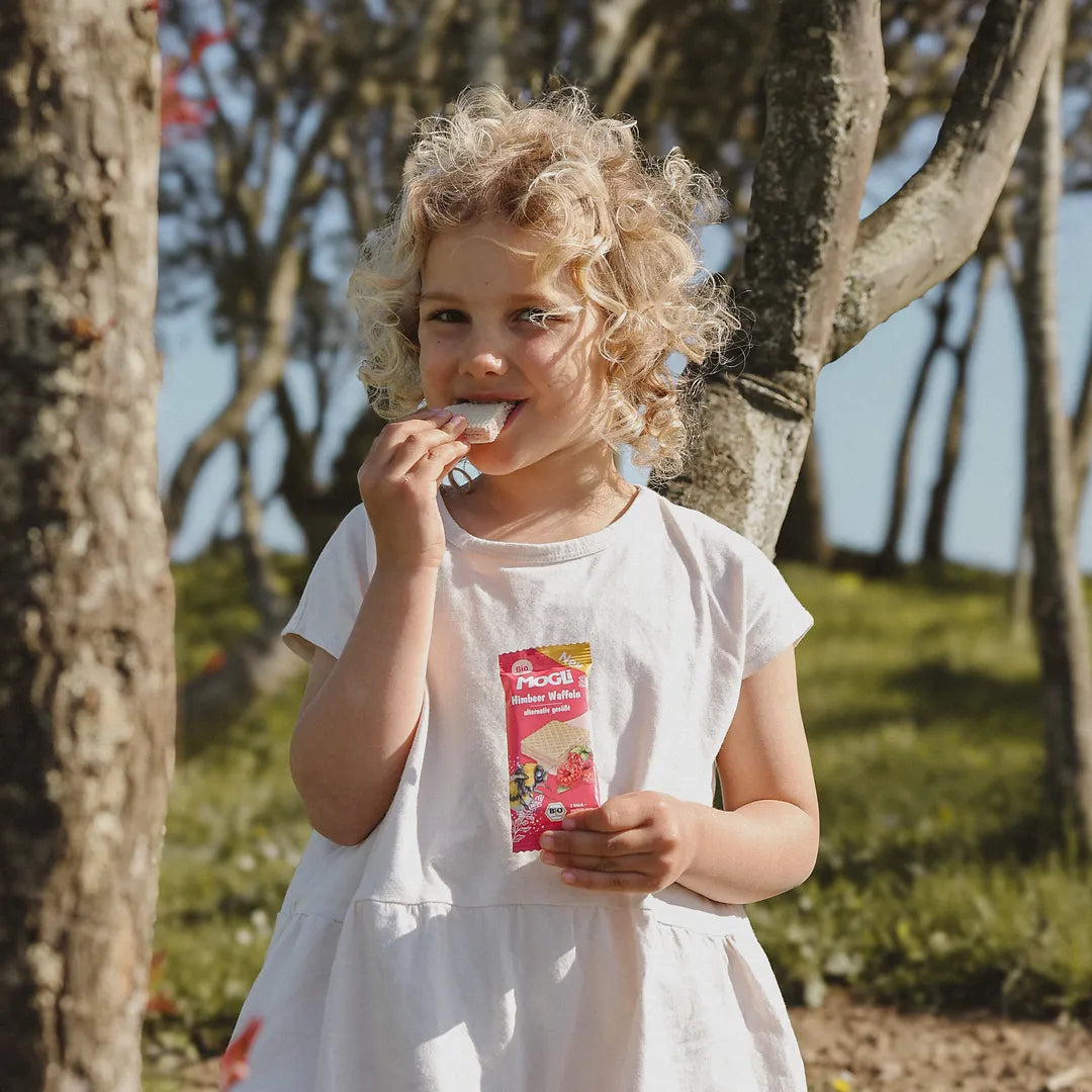 Kind isst MOGLi Himbeer Waffeln und hält die Verpackung in der Hand, aufgenommen in der Natur zwischen Bäumen.
