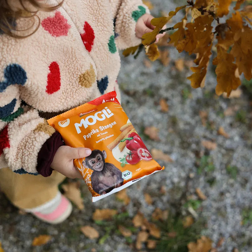 Kleines Kind hält eine Packung MOGLi Paprika Stangen in der Hand und in der anderen Hand einen Ast mit Herbstblättern