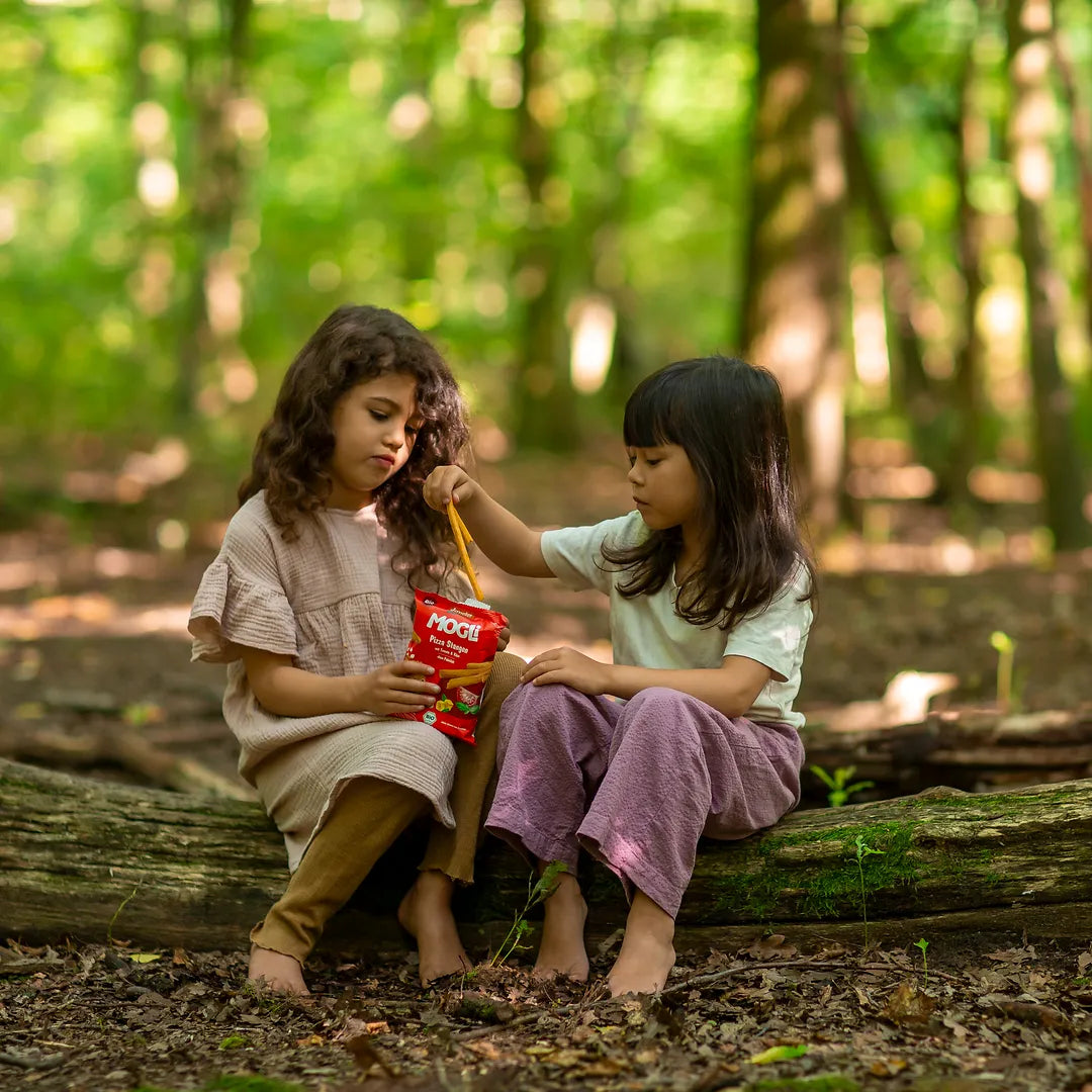 Zwei Mädchen im Wald sitzen auf einem Baumstamm und snacken die MOGLi Pizza Stangen