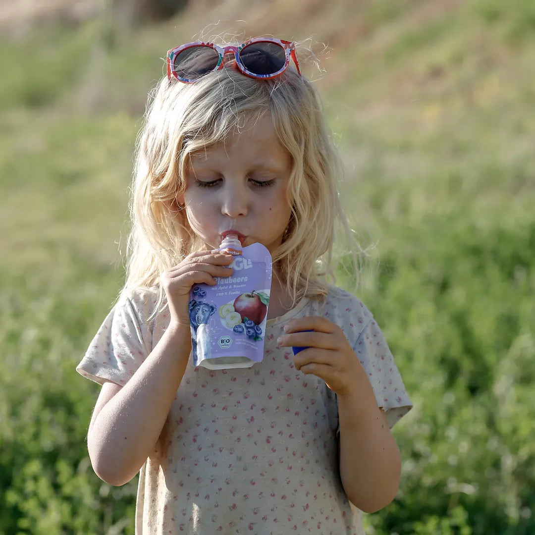 Mädchen trinkt MOGLi Blaubeer Quetschie auf einer Wiese.
