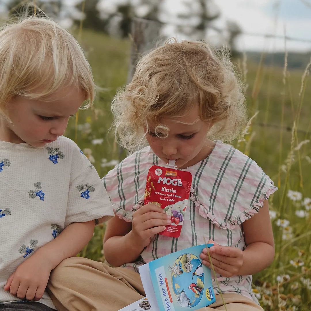Zwei Kinder schauen in ein Pixi Buch und ein Kind trinkt einen MOGLi Kirsch Quetschie.