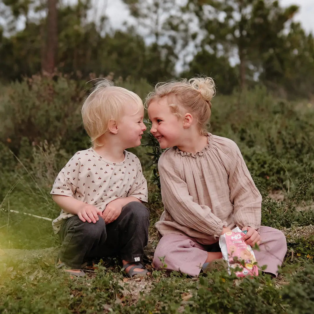 Zwei Kinder sitzen auf einer Wiese und snacken eine Packung MOGLi Zitronen Himbeer Kekse.