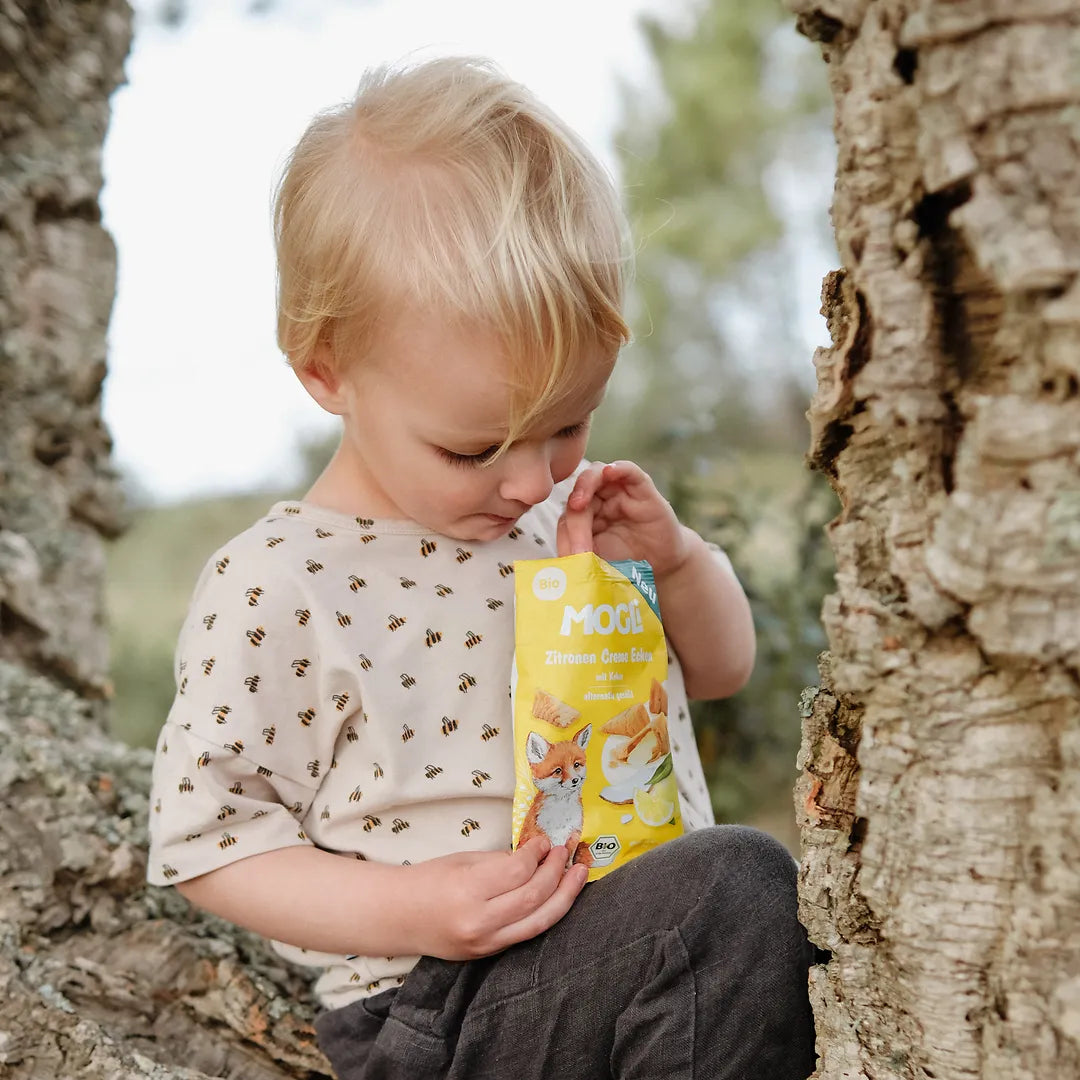 Blonder Junge sitzt auf einem Baum und schaut in eine Packung MOGLi Zitronen Creme Ecken.