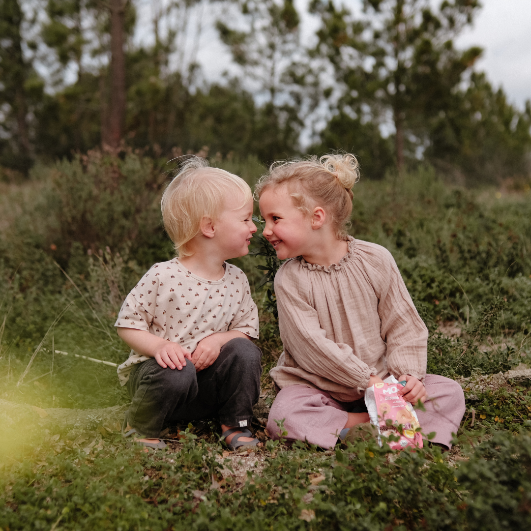 Blonder Junge und blondes Mädchen grinsen sich gegenseitig an und snacken die MOGLi Zitronen Himbeer Kekse in der Natur