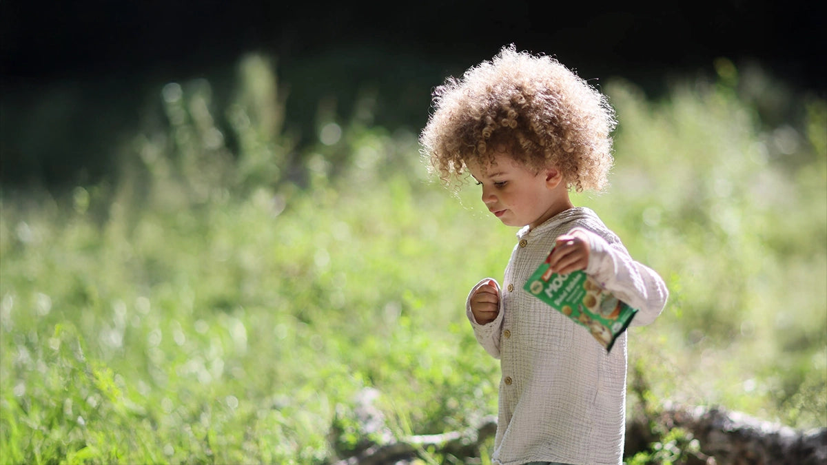Kleiner Junge mit Lockenkopf im Wald und einer Packung MOGLi Dinkel Bällchen Gartenkräuter in der Hand