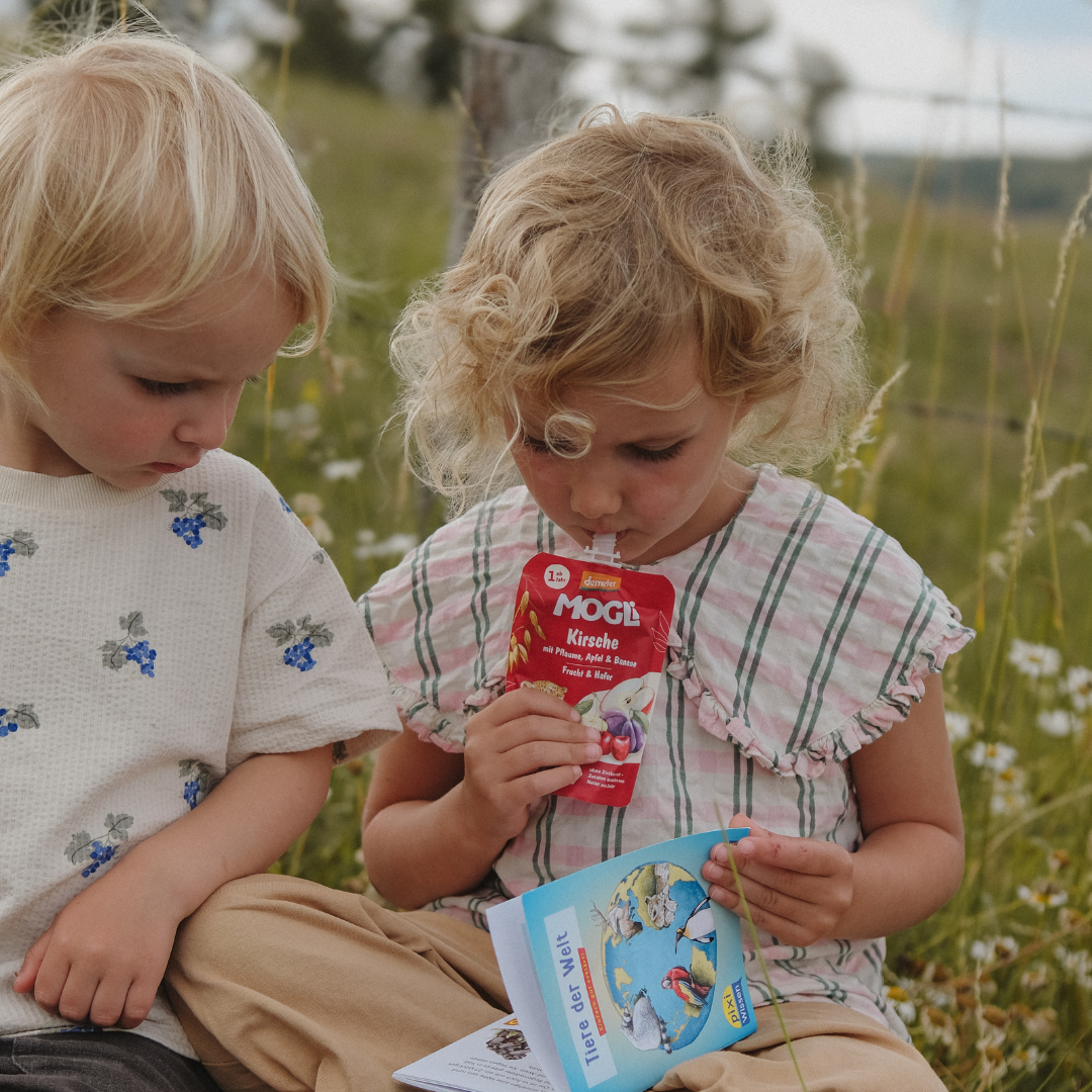 Junge und Mädchen schauen in ein Pixi-Buch mit dem Titel "Tiere der Welt" und das Mädchen trinkt den Quetschie Kirsche von MOGLi