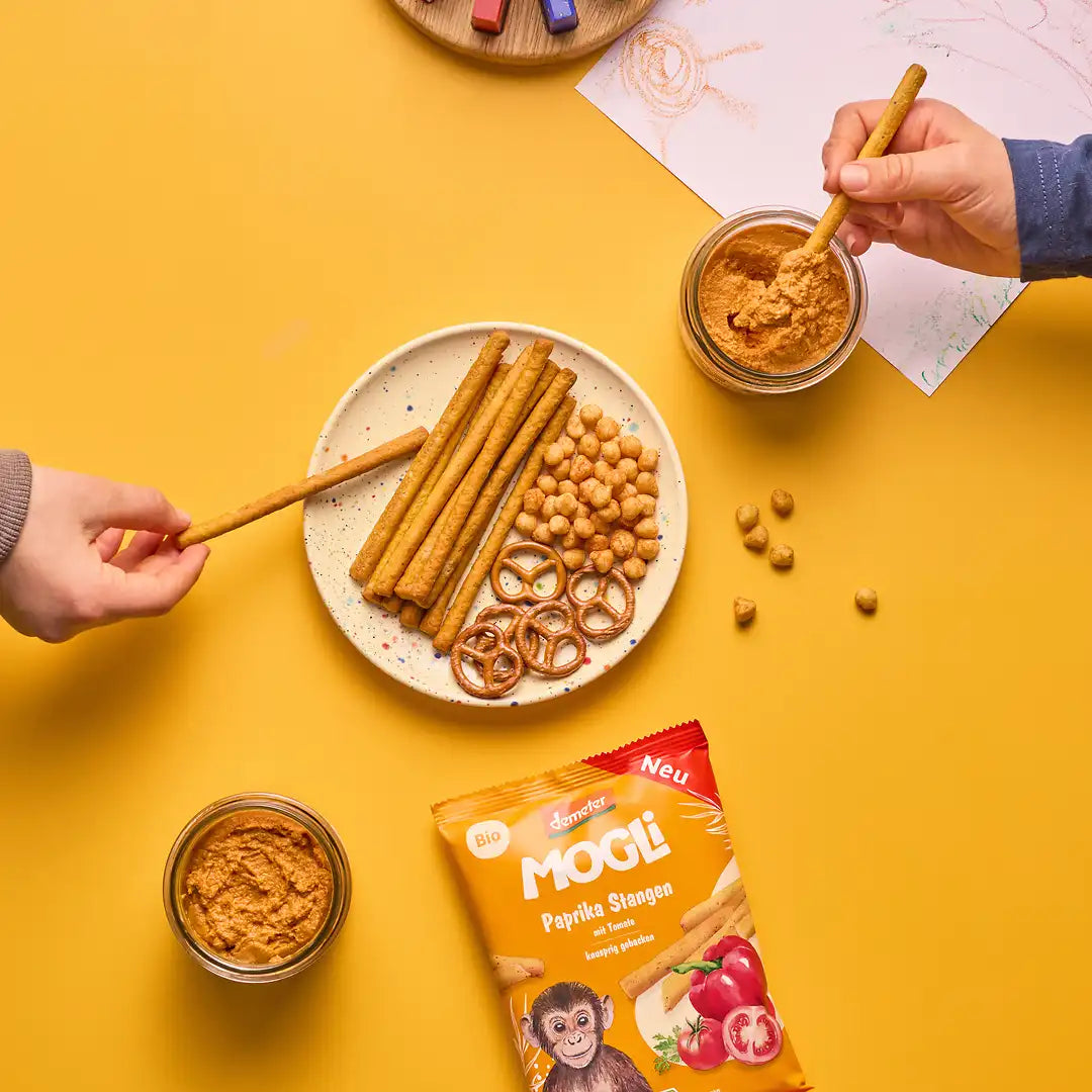 Zwei Kinder greifen auf einen Snackteller gefüllt mit herzhaften MOGLi Snacks und ein Kind dippt die Paprika Stange in den Zwergen Streich.