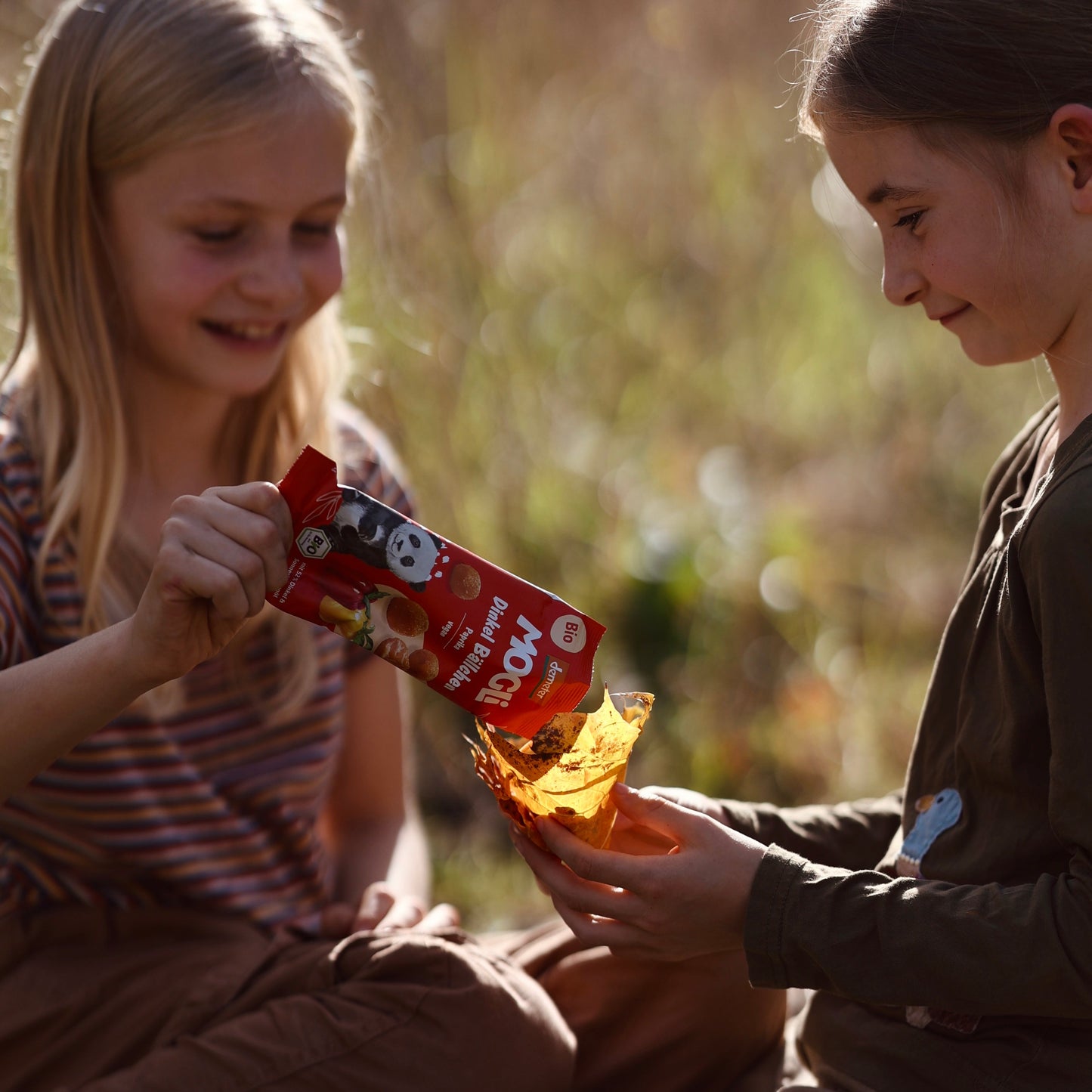 Zwei Mädchen in der Natur snacken die MOGLi Dinkel Bällchen Paprika, ein Mädchen füllt die Bällchen in ein Blatt, was als Schüssel dient