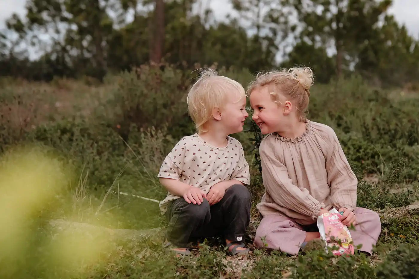 Blonder Junge und blondes Mädchen grinsen sich gegenseitig an und snacken die MOGLi Zitronen Himbeer Kekse in der Natur
