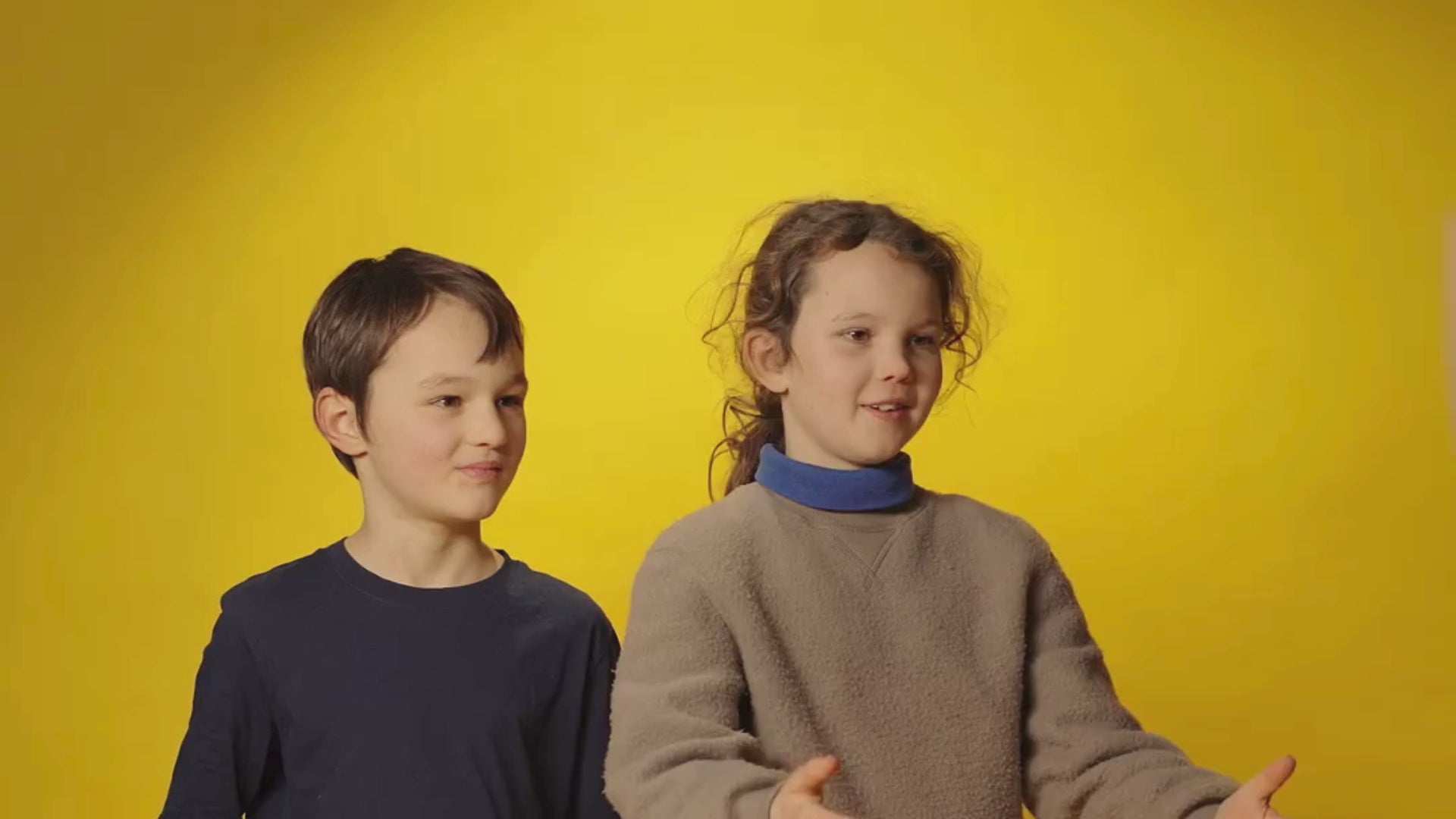 Zwei Kinder stehen vor einer gelben Wand und packen ein MOGLi Snackpaket aus. Das Paket ist gefüllt mit herzhaften Snacks und einem Zwergen Streich von Zwergenwiese.